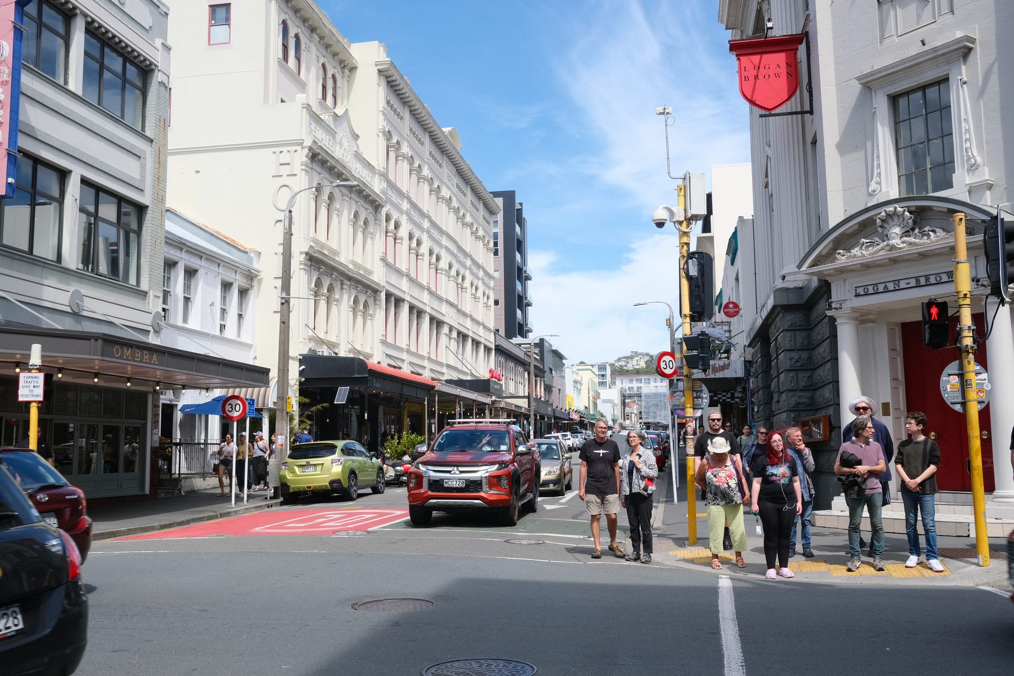 20 people struggle to fit on the footpath on Cuba street while cars go past. 
